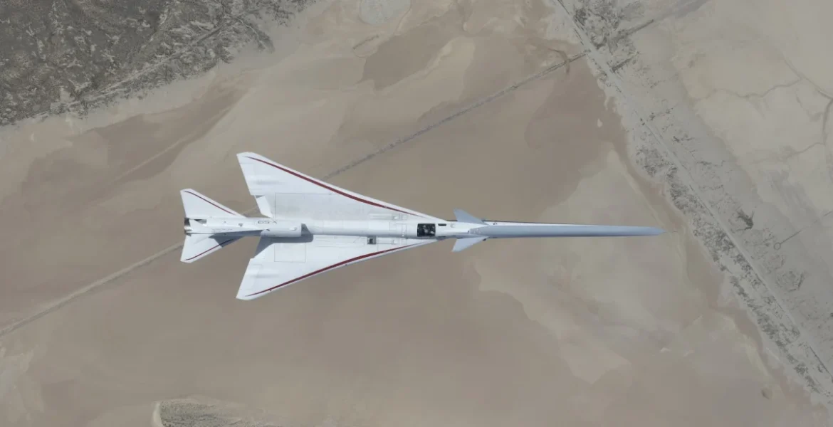 NASA’s white, blue, and red X-59 research aircraft flies over the Mojave Desert near NASA’s Armstrong Flight Research Center in Edwards, California, during its third flight. The aircraft is centered in the frame, viewed from above with the canopy, both wings, NASA and X-59 logos visible, and the landing gear down with one wheel visible. Below, rugged terrain transitions into the smooth texture of a dry lakebed as the aircraft, nearly 100 feet long, flies toward the right side of the frame.